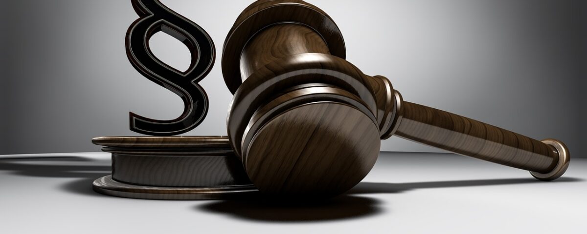 Ein hölzerner Richterhammer liegt neben einem Podest, auf dem ein großes Paragrafenzeichen steht – Symbol für Recht, Gesetz und gerichtliche Entscheidungen.