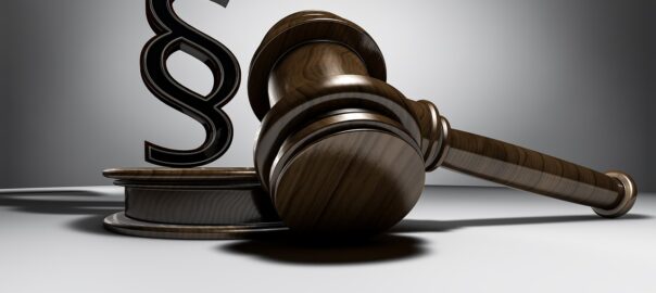 Ein hölzerner Richterhammer liegt neben einem Podest, auf dem ein großes Paragrafenzeichen steht – Symbol für Recht, Gesetz und gerichtliche Entscheidungen.