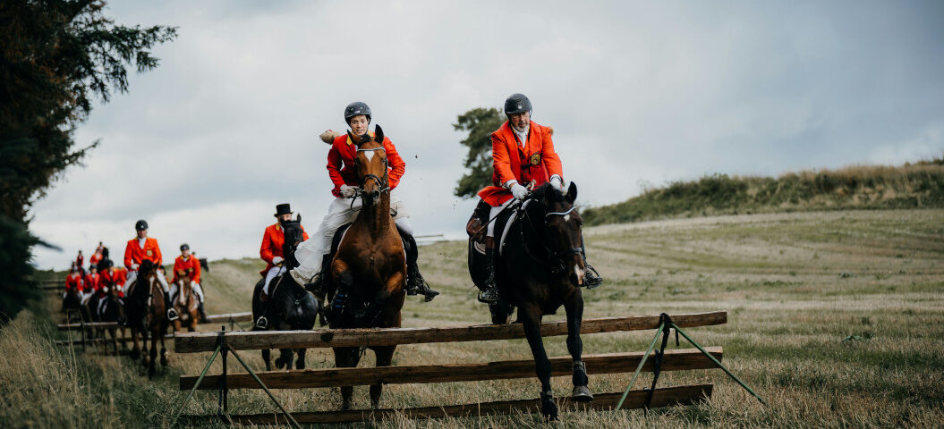 Reiter in traditionellen orangefarbenen Jagdjacken galoppieren bei einer Schleppjagd über eine Wiese und überspringen ein Holzhindernis.