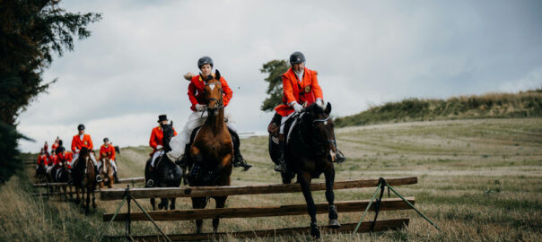 Reiter in traditionellen orangefarbenen Jagdjacken galoppieren bei einer Schleppjagd über eine Wiese und überspringen ein Holzhindernis.