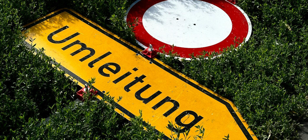 Gelbes Umleitungsschild und ein Durchfahrt-verboten-Schild liegen umgekippt im Gebüsch.