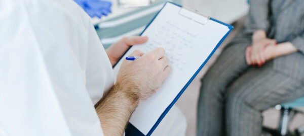 Ein Arzt dokumentiert während eines Patientengesprächs Notizen auf einem Klemmbrett.
